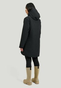 Svart vattentålig kappa med huva, långa ärmar och en avslappnad passform, som matchas med beige gummistövlar som når mitt på vaden och svarta leggings.