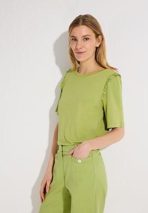 Frau trägt ein hellgrünes Kurzarmshirt mit Rüschen und passende hoch taillierte Hosen, steht vor einem weißen Hintergrund.