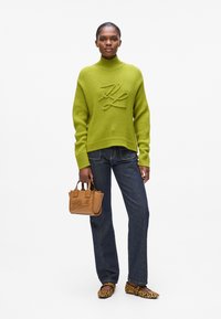 Grüner gerippter Pullover mit Rollkragen und geprägt Design, kombiniert mit dunkler Jeans und einer hellbraunen Handtasche, ergänzt durch Ballerinas im Leopardenmuster.