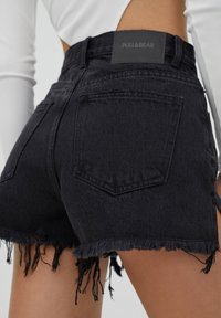 Pantalones cortos de mezclilla negra con dobladillo deshilachado, cintura alta, dos bolsillos traseros y una etiqueta de marca en la cinturilla trasera.