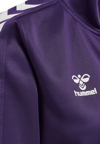 Primo piano di giacca sportiva viola con strisce bianche a chevron sulla spalla e logo bianco dell'ape hummel sul petto.