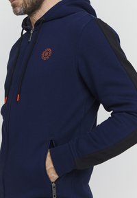 Marineblaue Kapuzenjacke mit schwarzen Akzenten. Verfügt über eine Kapuze mit Kordelzug, seitliche Taschen und ein kleines oranges Logo auf der Brust. Weiches, strukturiertes Material.