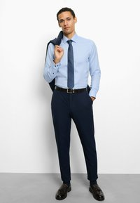 Light blue dress shirt, navy trousers, patterned tie, black belt, and shoes; jacket draped over one shoulder. Smooth fabric and tailored fit.