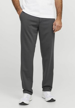 Homme portant un pantalon droit gris foncé, des baskets blanches et une chemise blanche, les mains détendues, une dans la poche, sur un fond uni.