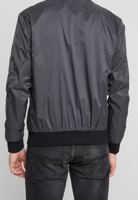 Svart rutigt bomberjacka med ribbade ärmslut och fåll, med en slät textur och en figursydd silhuett. Lämplig för avslappnad stil.