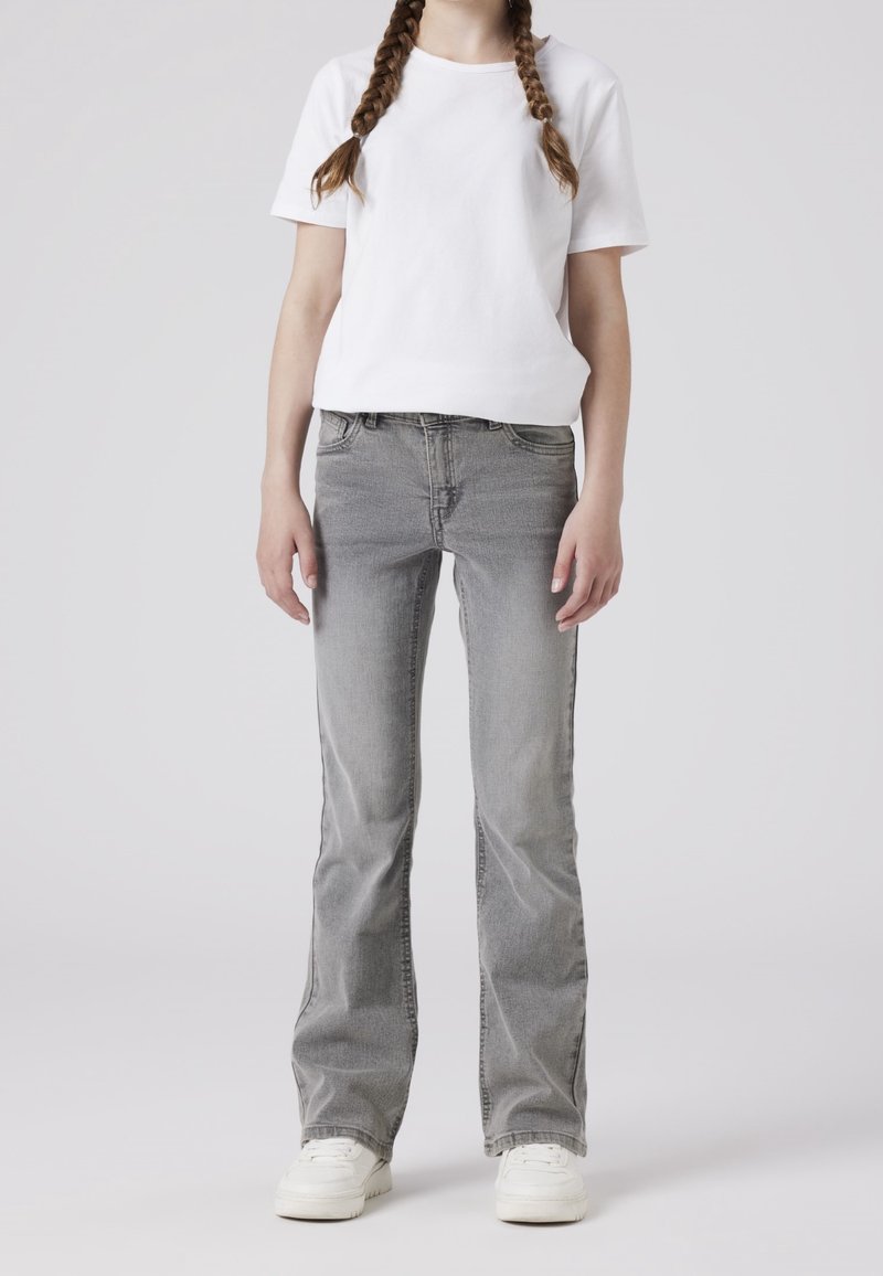 Personne portant une chemise blanche à manches courtes, un jean gris, des baskets blanches, avec deux longues nattes tressées, debout devant un fond uni.