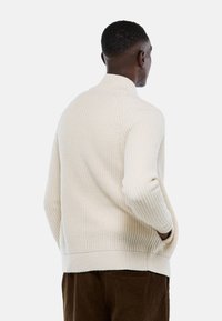 Pull en maille couleur crème avec un col montant, texture côtelée et épaules tombantes, associé à un pantalon en velours côtelé marron.