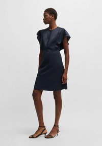 Mörkblå ärmlös klänning med volangaxlar, figurnära överdel och utsvängd kjol, tillverkad av slät tyg, tillsammans med strapatsandaletter.