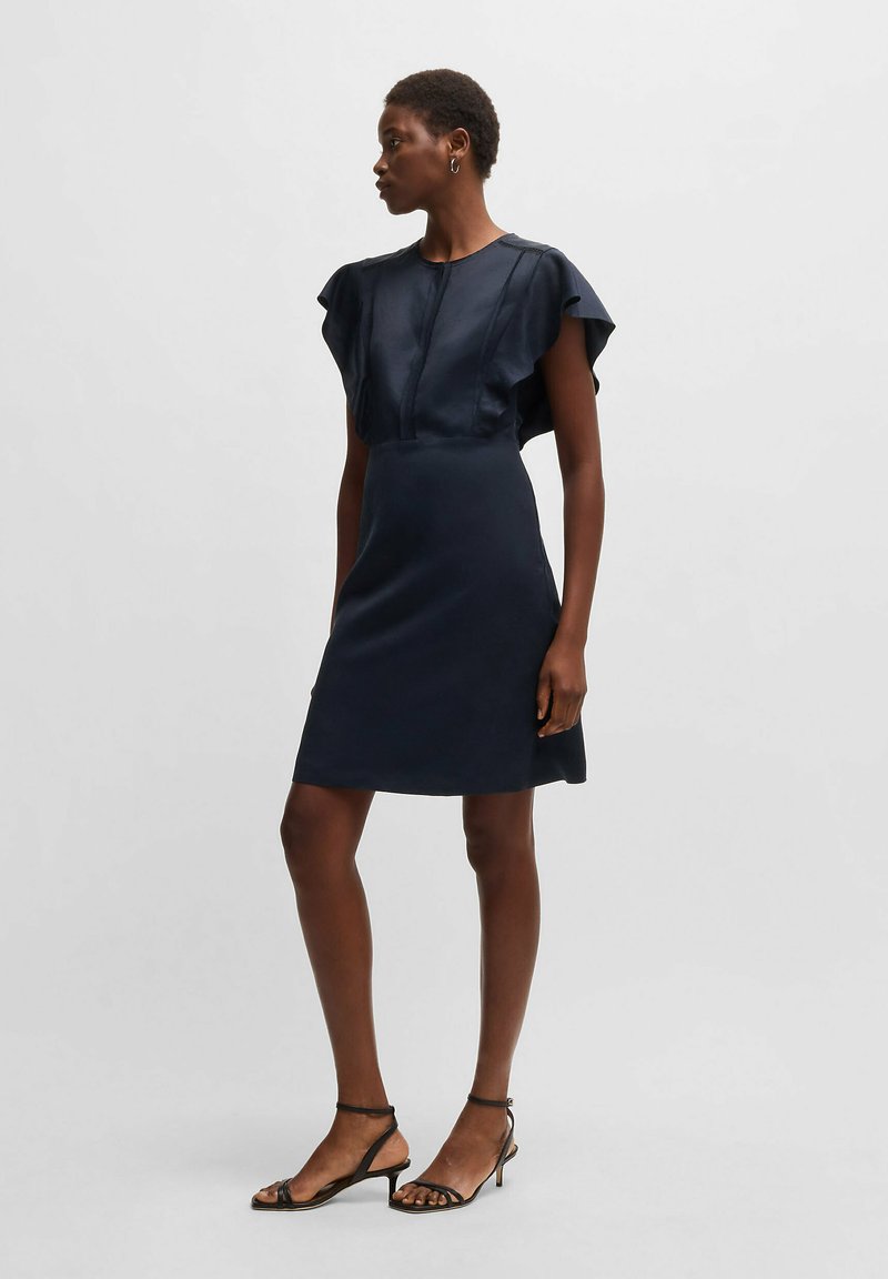 Mörkblå ärmlös klänning med volangaxlar, figurnära överdel och utsvängd kjol, tillverkad av slät tyg, tillsammans med strapatsandaletter.