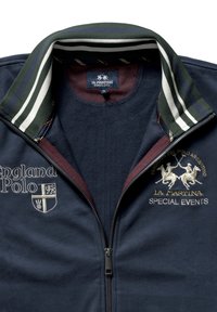 Mörkblå zip-up-jacka med grön och vit randig krage. Har broderade logotyper och text, texturerat tyg och dragkedja.