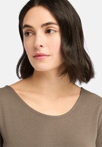 Braun, figurbetontes Oberteil mit weitem Rundhalsausschnitt. Glatter, leichter Stoff; kurzes, leicht welliges Haar ist an den Rändern sichtbar.
