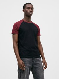 Giovane uomo che indossa una maglietta raglan nera e bordeaux e jeans cargo grigi, in piedi davanti a uno sfondo chiaro e semplice.
