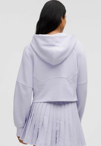Sudadera corta de color lila con capucha, que presenta costuras texturizadas y un logo en la parte posterior, combinada con una falda plisada a juego.