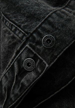 Tessuto in denim nero con finitura testurizzata, dotato di due bottoni a pressione in metallo incisi con la dicitura "DENIM" per la chiusura su un design a strati.