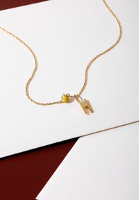 Goldene Kettenhalskette mit einem gelben Herzanhänger und einem goldenen Buchstaben "H" Anhänger. Der Hintergrund ist weiß, mit sichtbarem strukturiertem Papier.