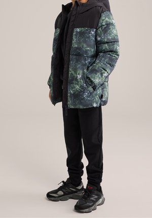 Doudoune noire avec un motif abstrait vert et bleu, dotée d'une capuche, d'une fermeture éclair à l'avant et de poches latérales. Associée à un pantalon de survêtement noir et des baskets.