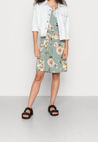 Robe fleurie verte avec des fleurs beige et orange, associée à une légère veste en denim. Des sandales noires à deux brides complètent le look.