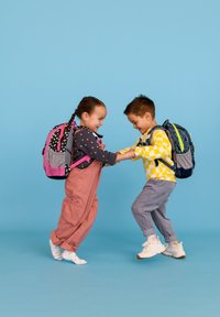 Zwei Kinder halten Händchen und tragen bunte Rucksäcke. Eines hat einen pinkfarbenen Rucksack mit Punkten; das andere trägt einen blauen Rucksack mit grünen Akzenten.