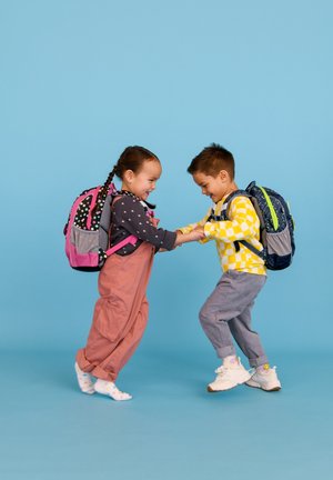 Due bambini si tengono per mano, indossando zainetti colorati. Uno ha uno zaino rosa con pois; l'altro indossa uno zaino blu con dettagli verdi.