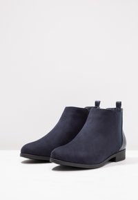 Stivali alla caviglia in suede blu navy con una texture liscia, pannelli laterali elastici e un tacco basso a blocco. Presentano un motivo a contrasto sul retro.