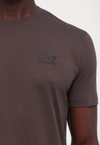 Tricou scurt, de culoare maro, fabricat din bumbac moale, având un logo negru EA7 Emporio Armani pe piept. Textură netedă, croială clasică.