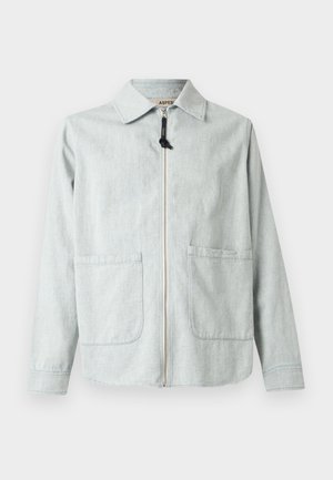 Vīrietis valkā gaiši pelēku jaku virs tumši zila T-krekla, pelēkas kravas bikses un baltus sporta apavus, stāvot pret vienkrāsainu balto fonu.