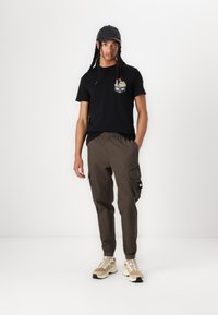 Black short-sleeve t-shirt with graphic detail on the chest, paired with olive-green cargo pants featuring side pockets, and beige sneakers.