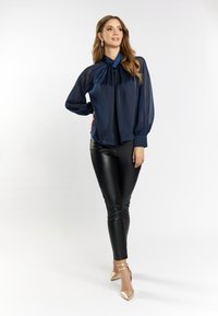 Mørkeblå, gennemsigtig bluse med høj krave og plisseret front, parret med sorte faux læder leggings og guld hæle.