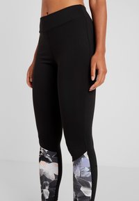 Kobieta w czarnych legginsach z kwiatowymi panelami na dolnych nogawkach, stojąca na jednolitym jasnym tle.