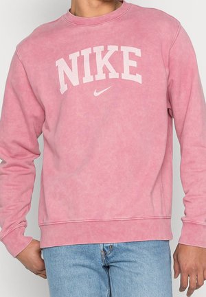 Person trägt einen ausgewaschenen rosa Nike-Sweatshirt mit weißem Logo und hellblauen Jeans, steht mit der linken Hand in der Tasche.