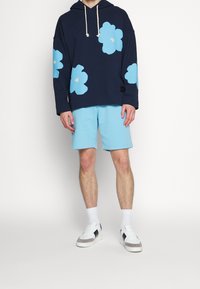 Felpa con cappuccio blu navy con grandi motivi floreali azzurri, abbinata a pantaloncini azzurri e sneaker bianche con dettagli grigi e strisce nere.