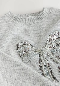 Jasnoszary sweter z dzianiny z dużą, srebrną cekinową kokardą przyszytą z przodu, w pobliżu dekoltu.