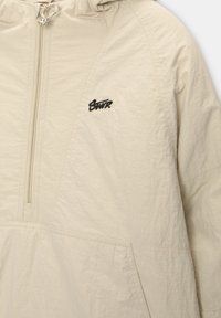 Veste beige à enfiler avec demi-fermeture éclair, capuche à cordon, poche avant et petit logo noir brodé sur la poitrine.