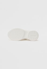 Semelle d'une basket blanche avec des motifs de grip texturés, vue par le dessous sur un fond blanc uni.