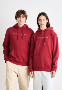 Sudaderas rojas con cordones, logo dorado "TOMMY JEANS", bolsillos frontales, textura suave. Combinadas con pantalones beige y azul claro para un contraste.