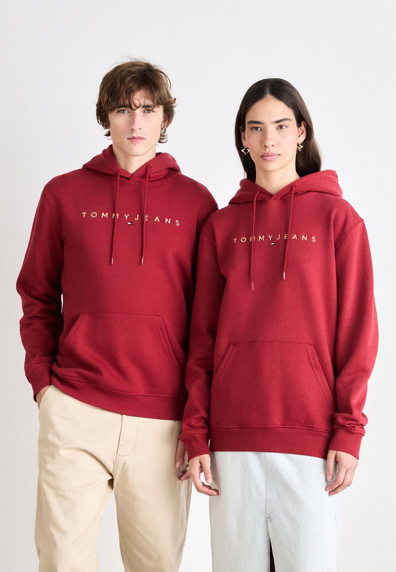 Sudaderas rojas con cordones, logo dorado "TOMMY JEANS", bolsillos frontales, textura suave. Combinadas con pantalones beige y azul claro para un contraste.