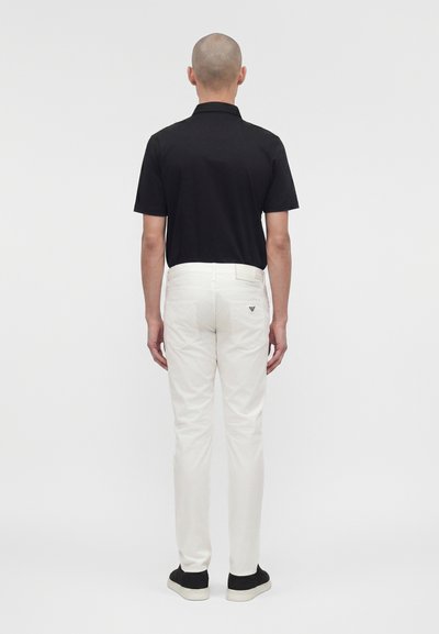 Άνδρας που φοράει μαύρο πουκάμισο με κοντά μανίκια, άσπρο παντελόνι με λογότυπο στην πίσω τσέπη και μαύρα παπούτσια, στέκεται με την πλάτη προς τα εμπρός σε μονόχρωμο φόντο.