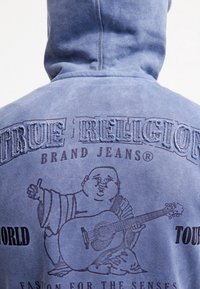 Felpa con cappuccio blu sbiadito con testo in rilievo "TRUE RELIGION BRAND JEANS" e grafica di una figura che suona una chitarra acustica.