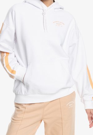 Personne portant un sweat à capuche blanc avec des rayures beige et orange sur les manches et un pantalon beige, tous deux avec le texte « ROXY Active by Nature ».