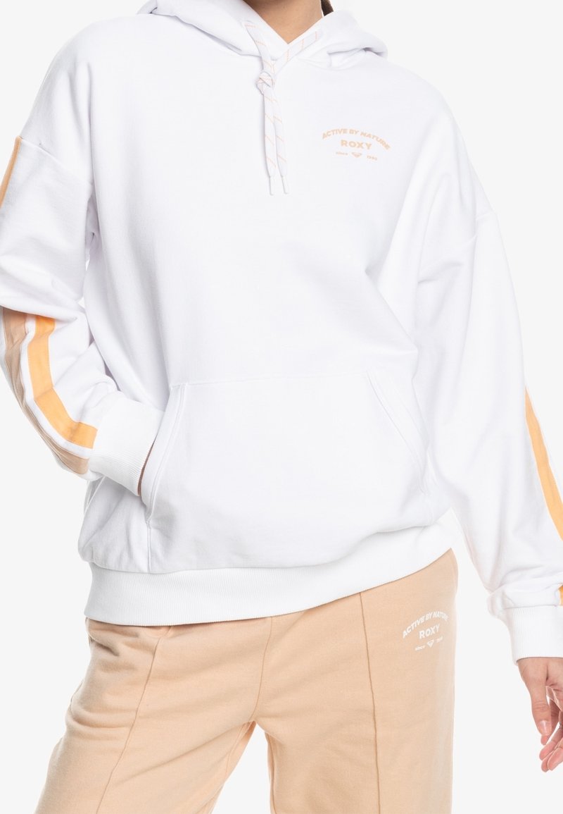 Personne portant un sweat à capuche blanc avec des rayures beige et orange sur les manches et un pantalon beige, tous deux avec le texte « ROXY Active by Nature ».