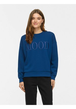 Jeune femme aux longs cheveux bruns portant un sweat-shirt bleu avec le mot "MOOD" brodé sur le devant et un pantalon noir.