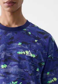 Blue patterned sweatshirt featuring a triangle logo, textured fabric with multicolored leaf-like accents. Crew neck and short sleeves.