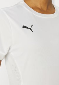 Hvit idrettskjorte laget av glatt stoff med rund halsutskjæring og svart Puma-logo på venstre bryst; har mesh-detaljer på sidene.