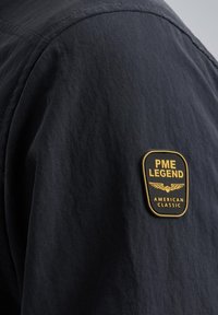 Schwarzer Stoff mit subtiler Textur, versehen mit einem rechteckigen gelb-schwarzen Patch, der mit "PME LEGEND AMERICAN CLASSIC" beschriftet ist.