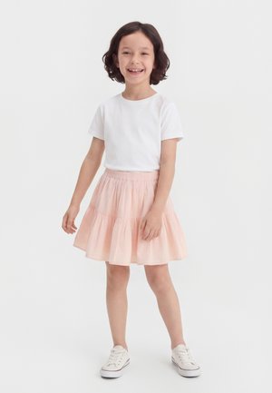 Nasmejan otrok s kratkimi kodrastimi lasmi stoji v beli majici, svetlo roza večplastni krilu in belih supergah na enobarvni podlagi.