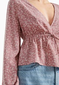 Blusa a fantasia floreale in morbido tessuto rosa pallido, con scollatura a V profonda, vita arricciata e maniche lunghe e larghe. Abbinata a pantaloni in denim.