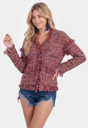 Kvinde iført en tekstureret rød og flerfarvet frynset cardigan med slidte denimshorts og blå kvastøreringe, smilende med hånd på hoften.