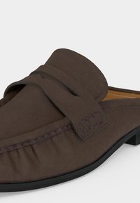 Mocassin marron en daim à enfiler avec bout arrondi, détails cousus et semelle noire sur fond blanc.