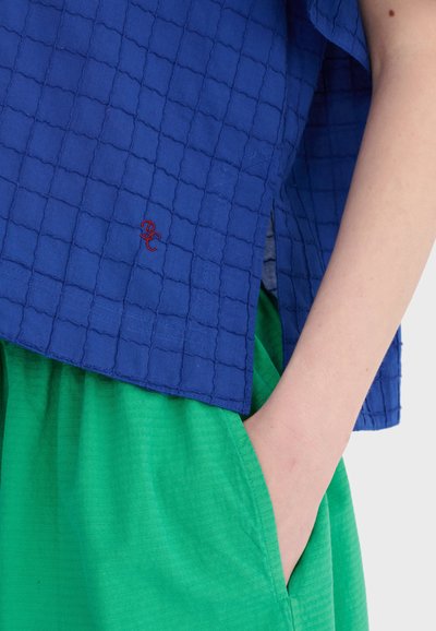 Osoba ubrana w niebieską, teksturowaną bluzkę z czerwonym logo oraz jasną zieloną spódnicę, z ręką swobodnie wsuniętą do kieszeni spódnicy, na jednolitym tle.