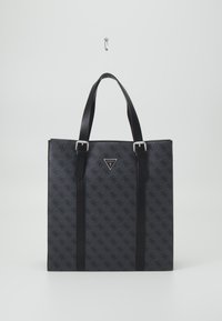 Svart Guess tote-väska med monogrammönster, silverfärjade spännen och dubbla axelremmar hängande mot en plain vit bakgrund.